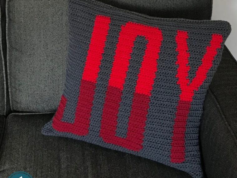 Crochet Pillow with the word joy in red sitting on a couch