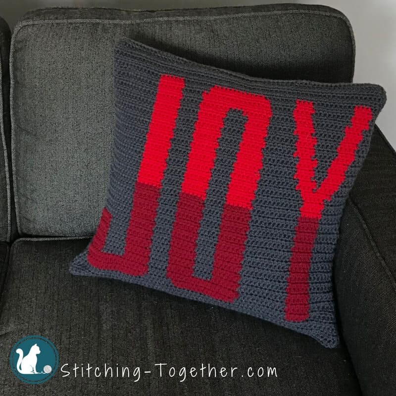 Crochet Pillow with the word joy in red sitting on a couch