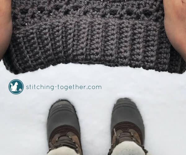 hands holding a crochet hat over the snow with boots visible