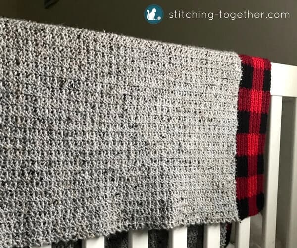 Gray baby blanket with buffalo plaid border draped on the side of a crib