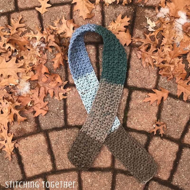 Brown and blue crochet boy scarf on the ground surrounded by fall leaves