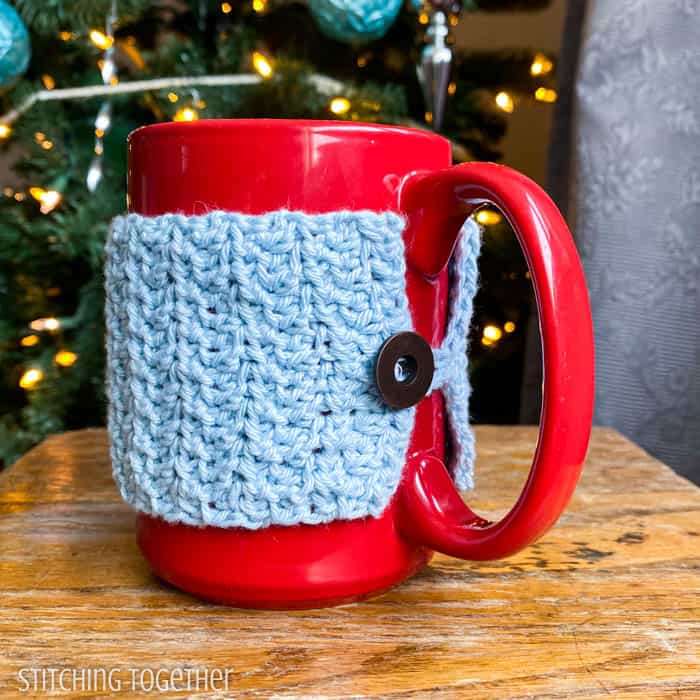 close up of crochet coffee mug cozy with button sitting on a down table