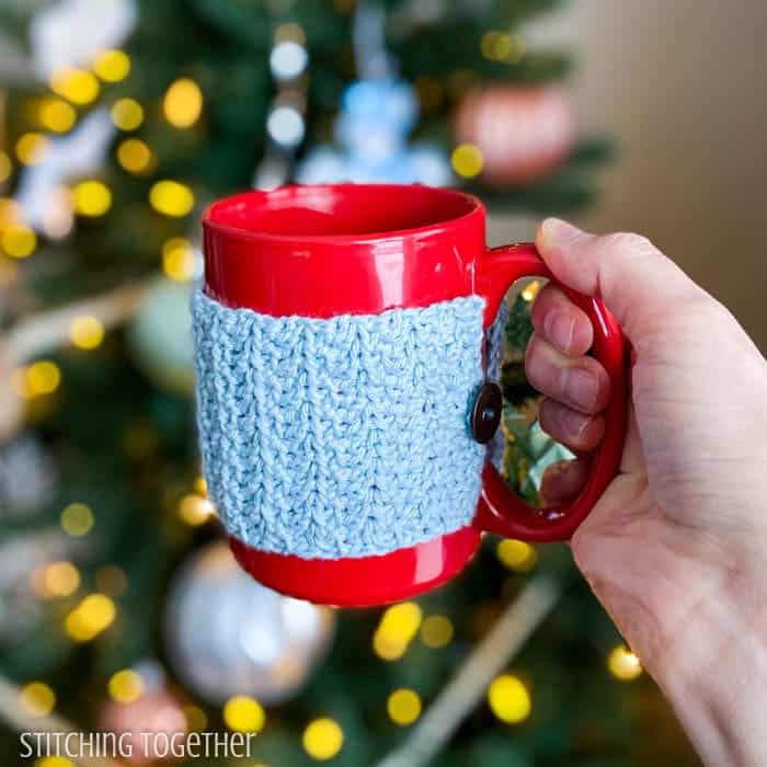 crochet mug cozy sitting on a brown table in front of a Christmas tree