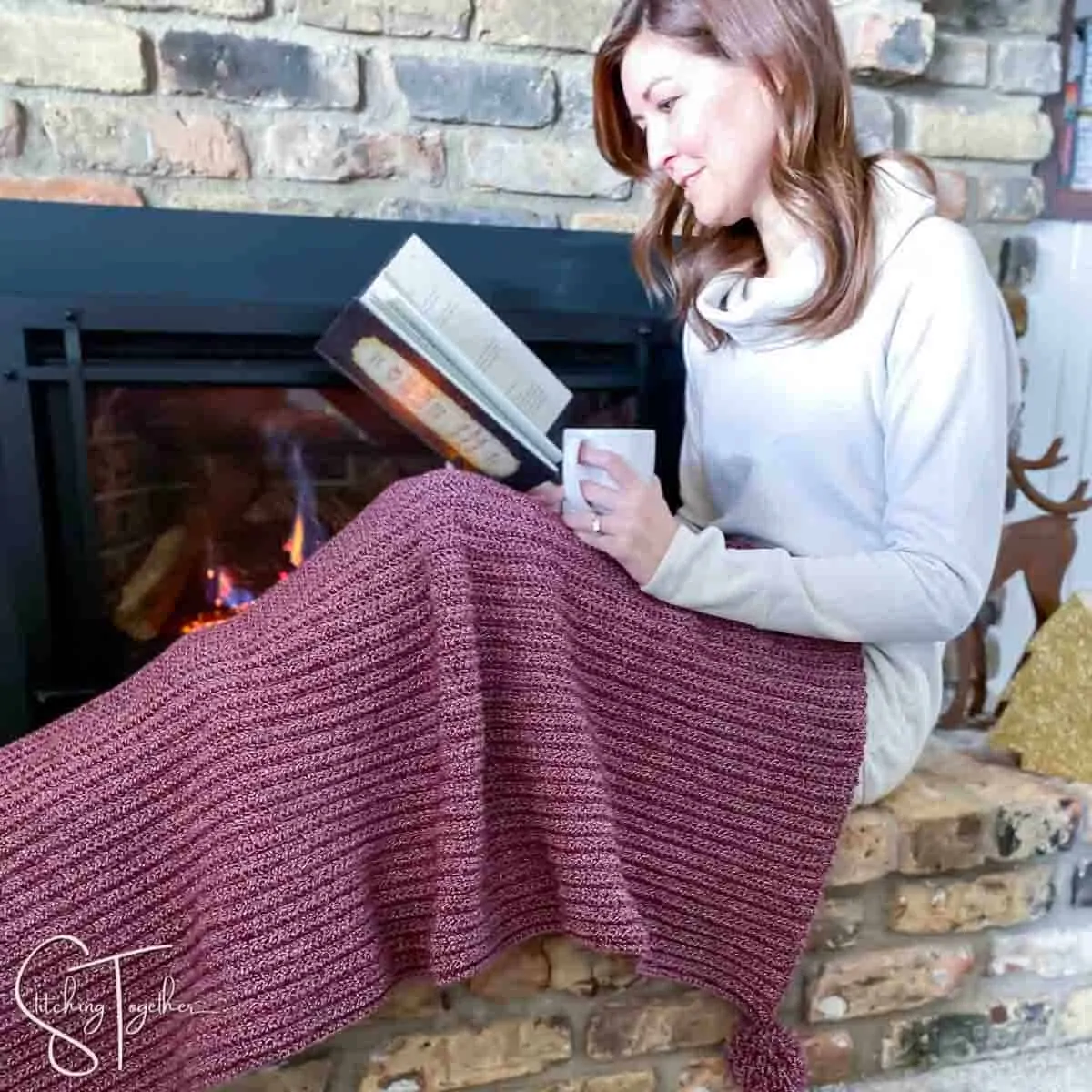 lady draped in a red and white crochet christmas blanket while reading in front of a fireplace