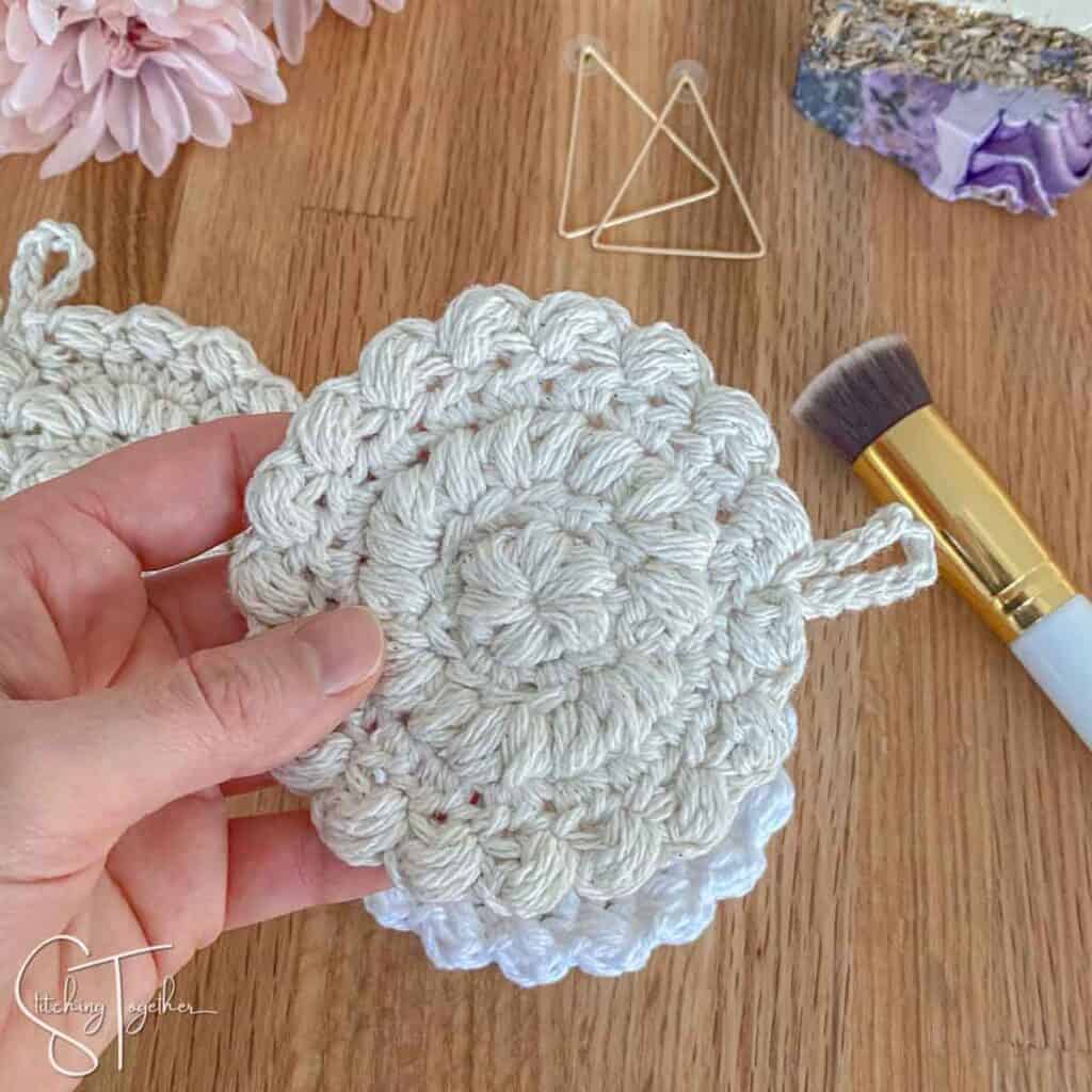a hand holding a crochet cotton face pad with accessories in the background