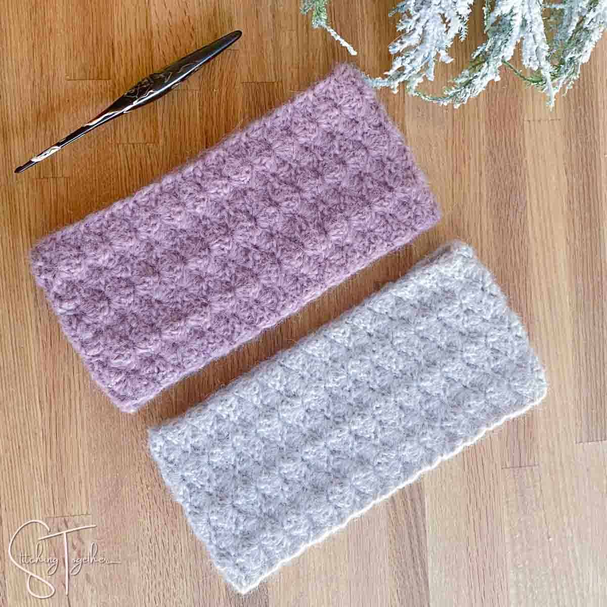 two shell stitch crochet ear warmers laying next to each other with a crochet hook and greenery