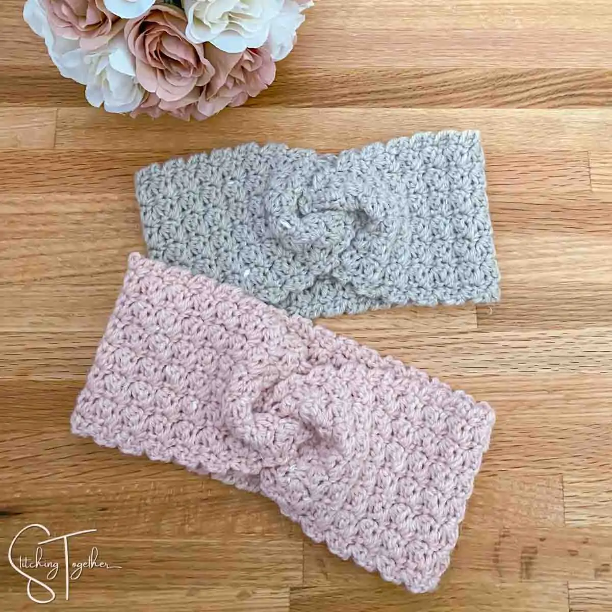 two twist crochet ear warmers laying flat next to a bouquet of roses