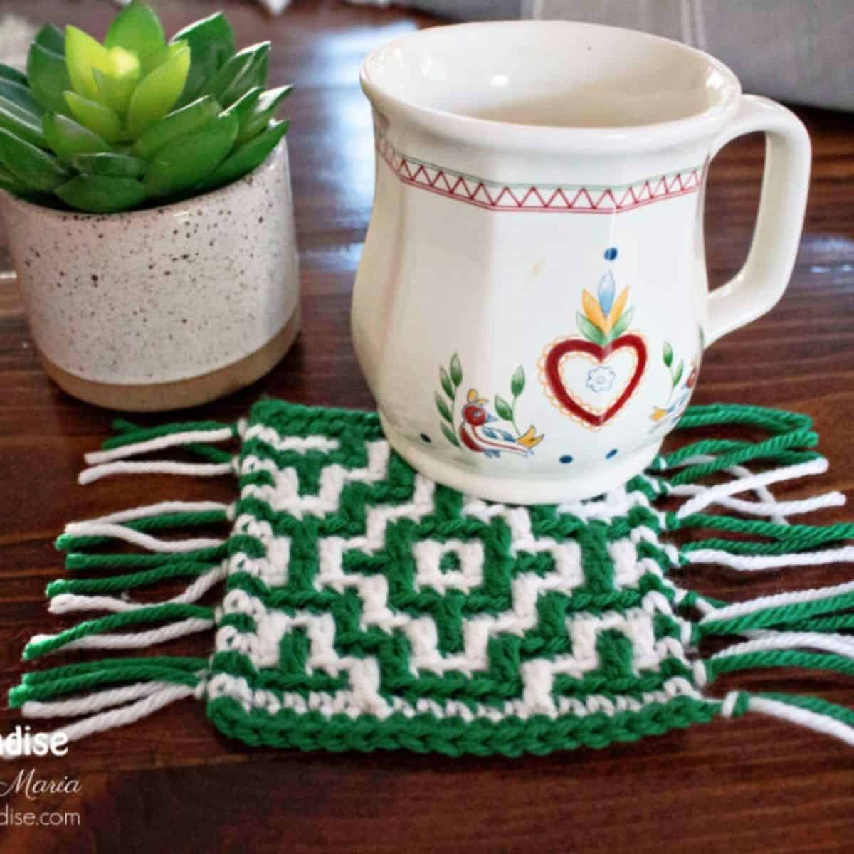 tea cup, plant, and mosaic green and white crochet coaster with fringe on a table