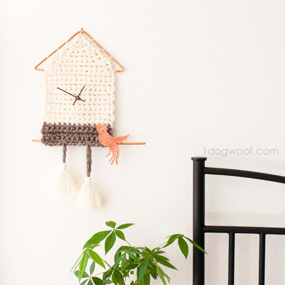 relógio de crochê fofo com um passarinho de crochê empoleirado nele pendurado ao lado de uma cama e acima de uma planta