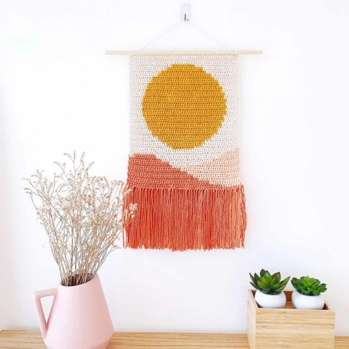 crochet tapestry with a sun over pink dunes hanging over a table top with plants