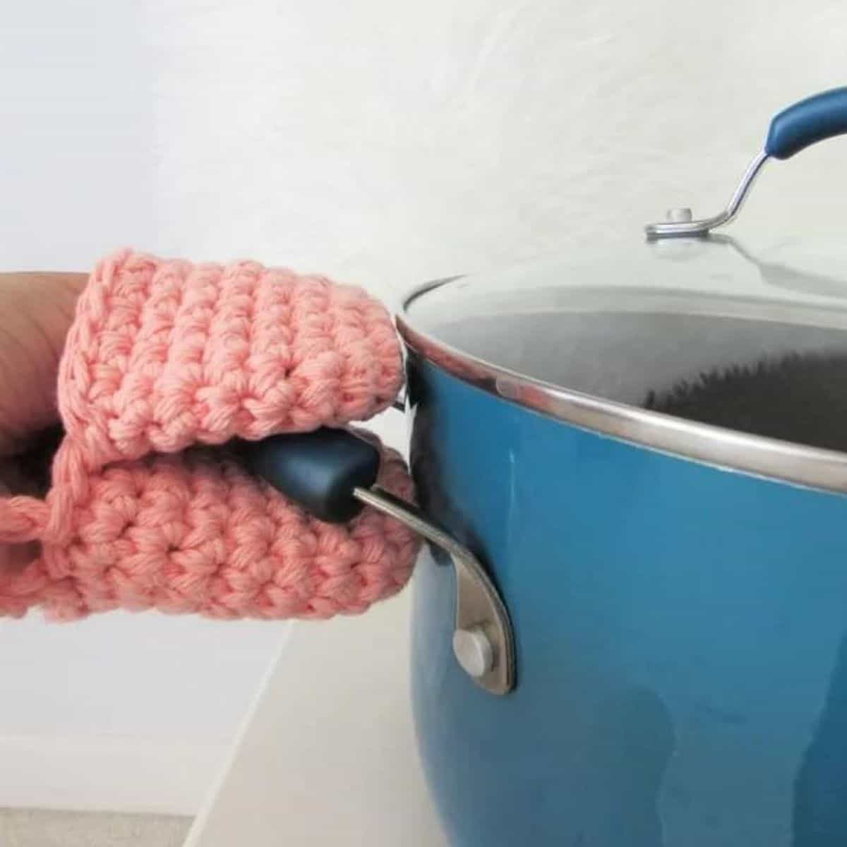 hand wearing a mini crochet oven mitt while grabbing the handle of a pot