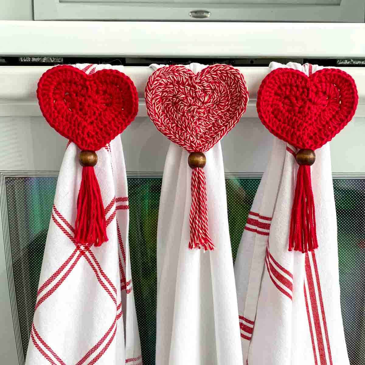 3 crochet heart towel toppers hanging 3 red and white towels on a stove handle