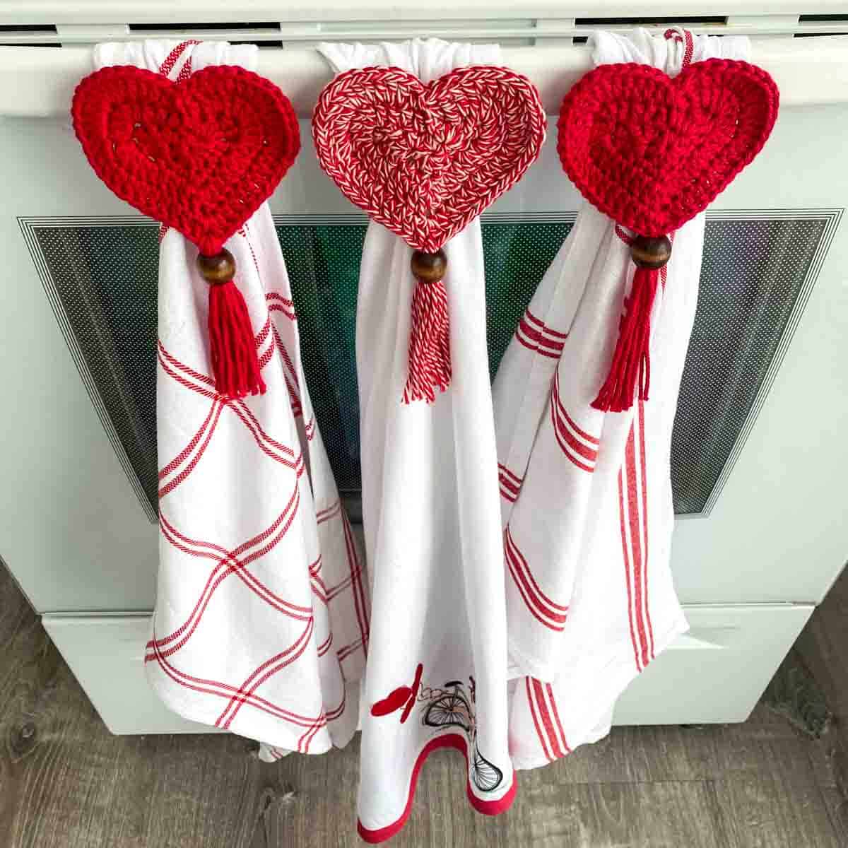3 Valentine dish towels with crochet heart toppers hanging on a oven