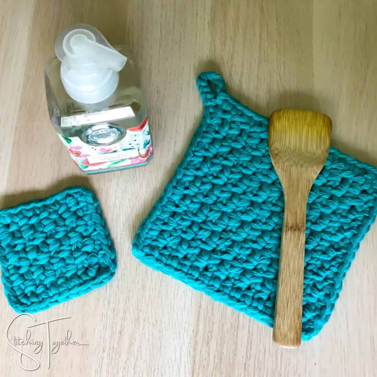 large and small crocheted pot holders laying flat with bottle of soap nearby and a wooden spoon laying on the large potholder