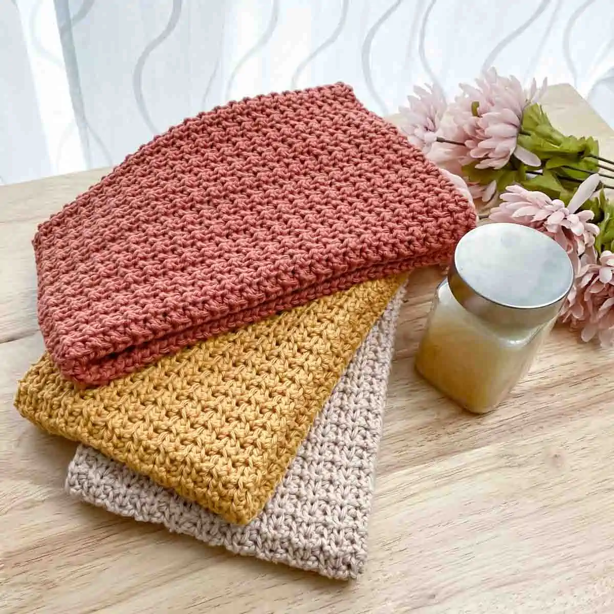 three crochet dish towels folded and loosely stacked next to some flowers and a jar