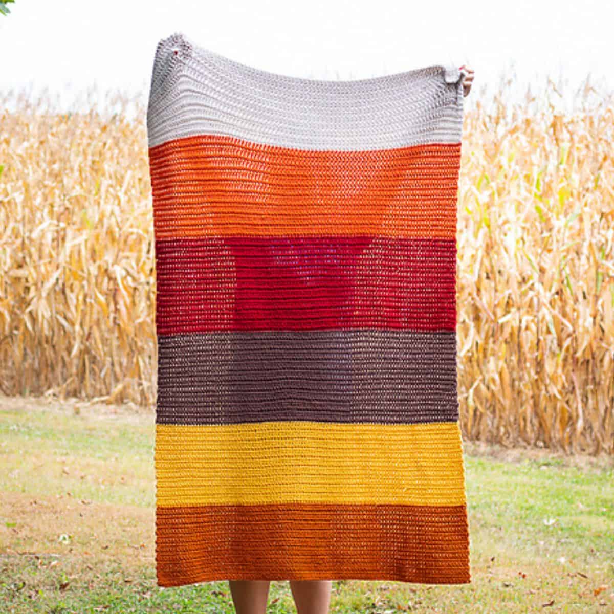 a striped crochet blanket in autumn colors being held up by a person in a field