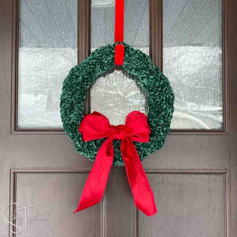 green crochet christmas wreath with a large red bow hanging on a front door by a red wreath hanger