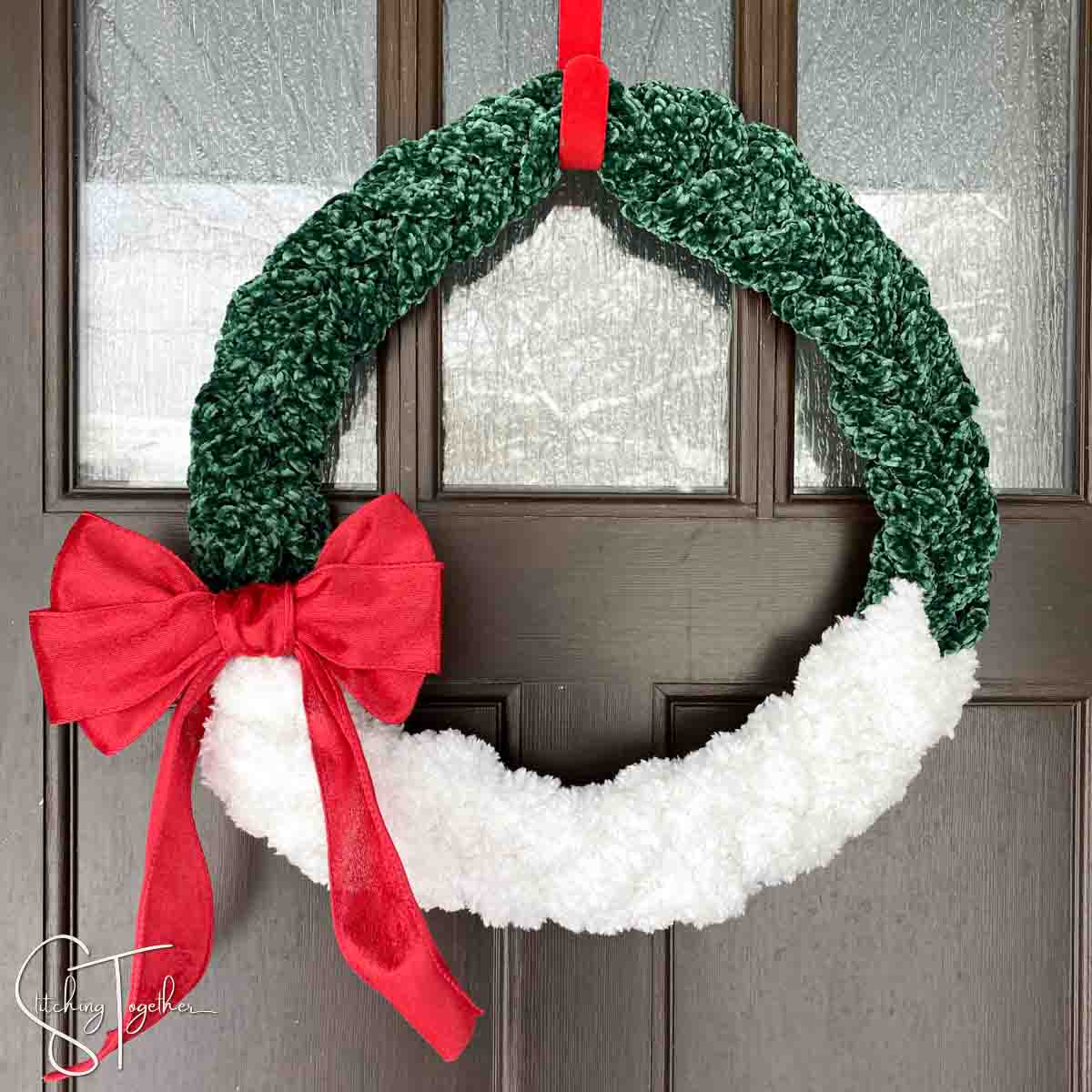 green and white crochet winter wreath with a large red bow hanging on a front door by a red wreath hanger