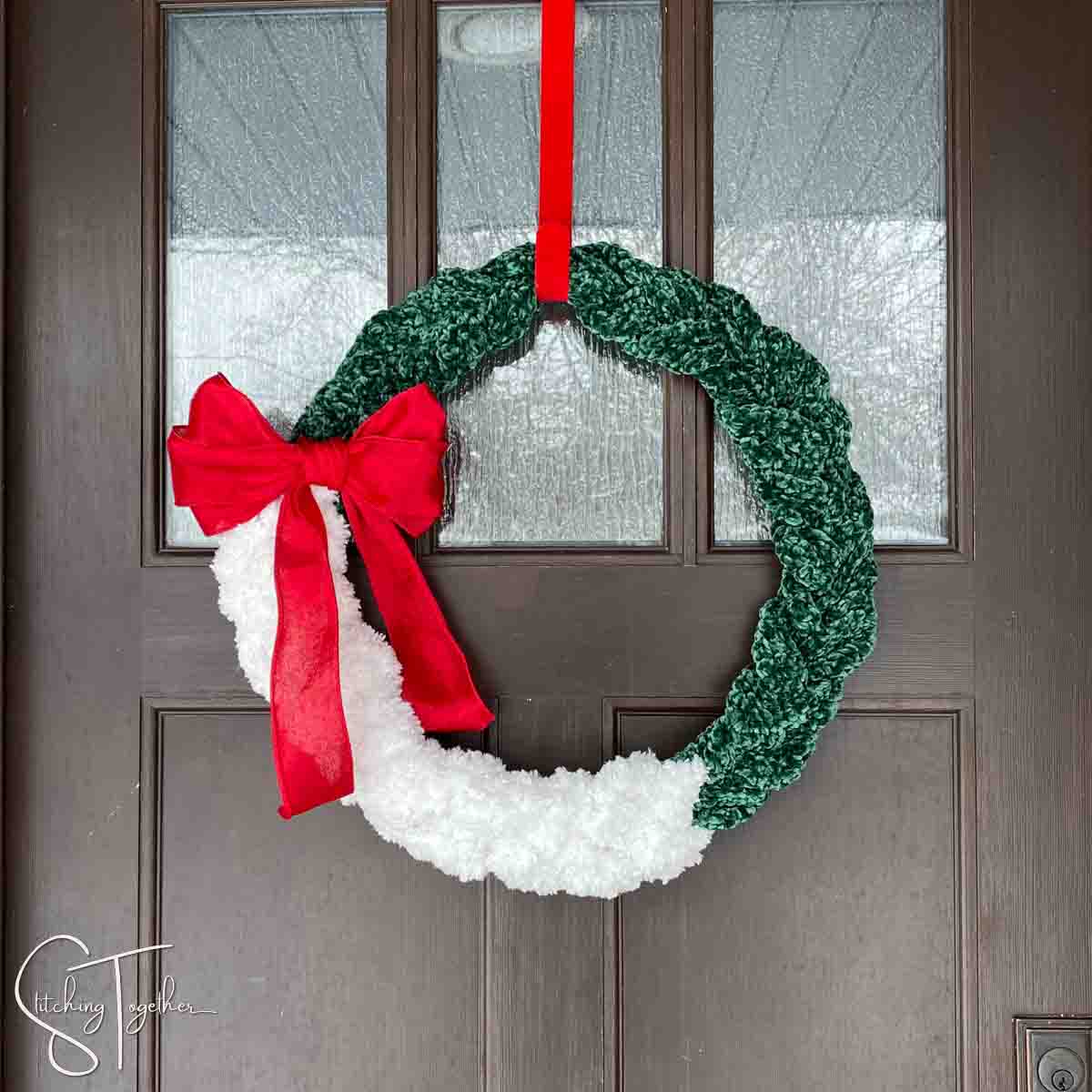 green and white winter crochet wreath with a large red bow hanging on a front door by a red wreath hanger