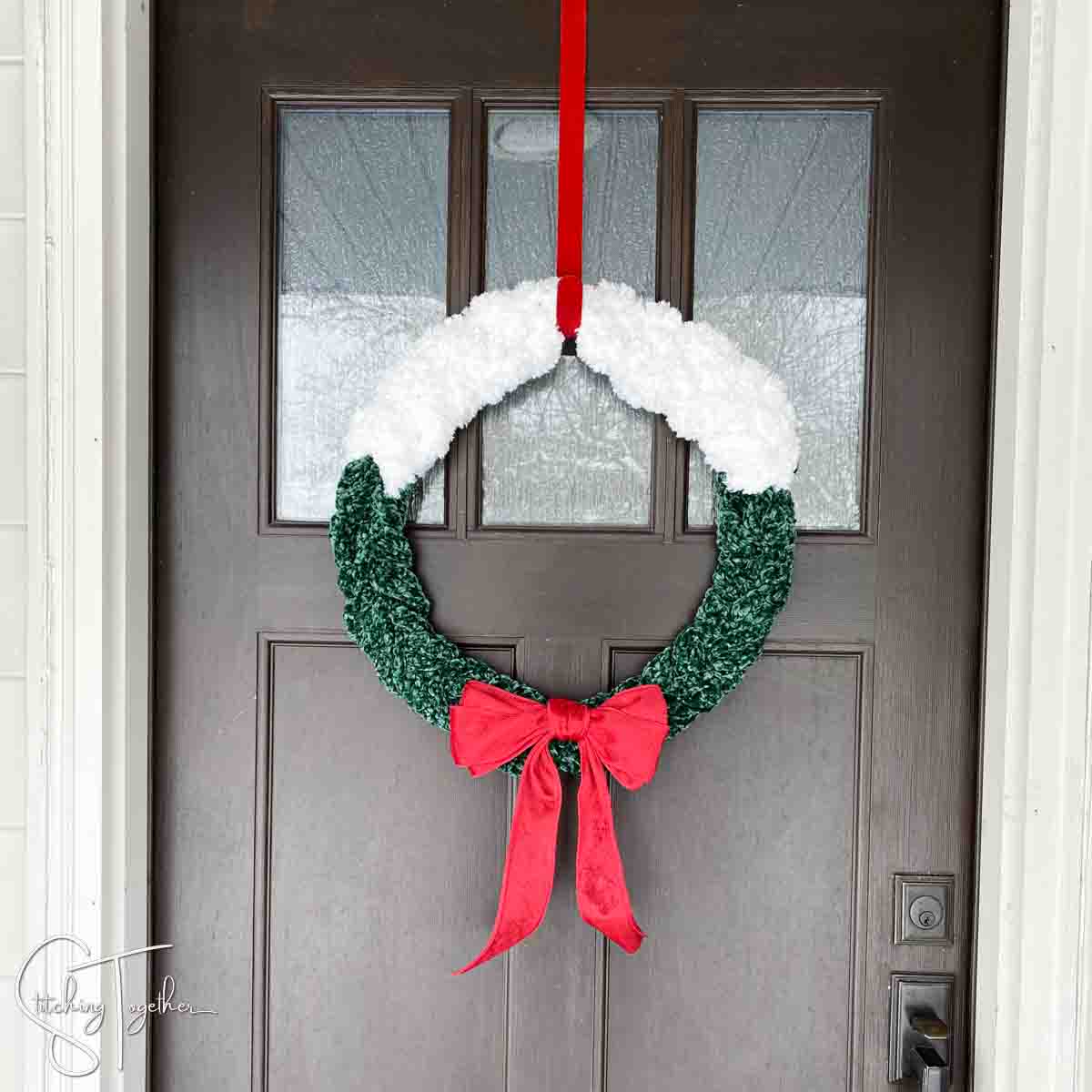 green and white Christmas crochet wreath with a large red bow hanging on a front door by a red wreath hanger