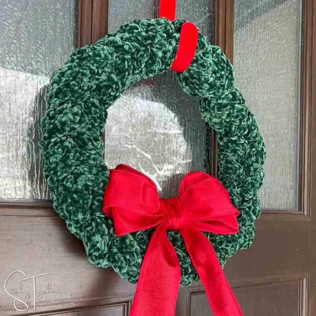 green Christmas crochet wreath with a large red bow hanging on a front door by a red wreath hanger