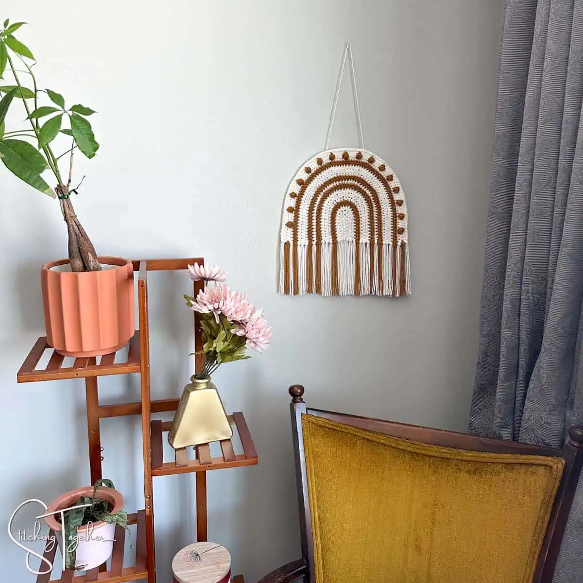 a boho crochet rainbow hanging with fringe on a wall next to a plant shelf and above a yellow chair