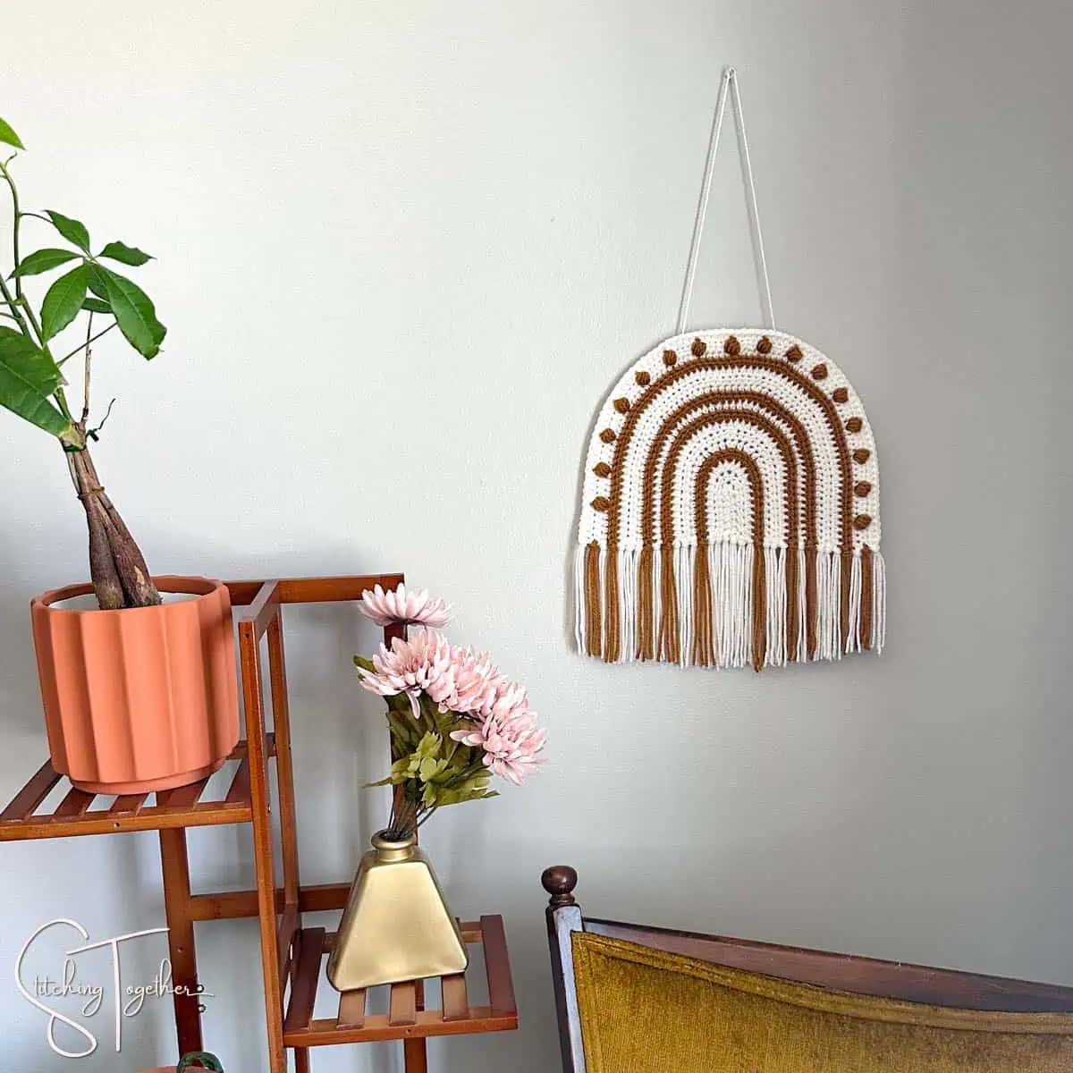 a boho rainbow crochet wall hanging with fringe on a wall next to a plant shelf and above a yellow chair