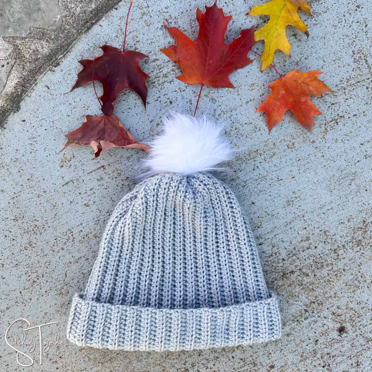 gray crochet ribbed hat with a white pom pom laying on the ground with colorful leaves around