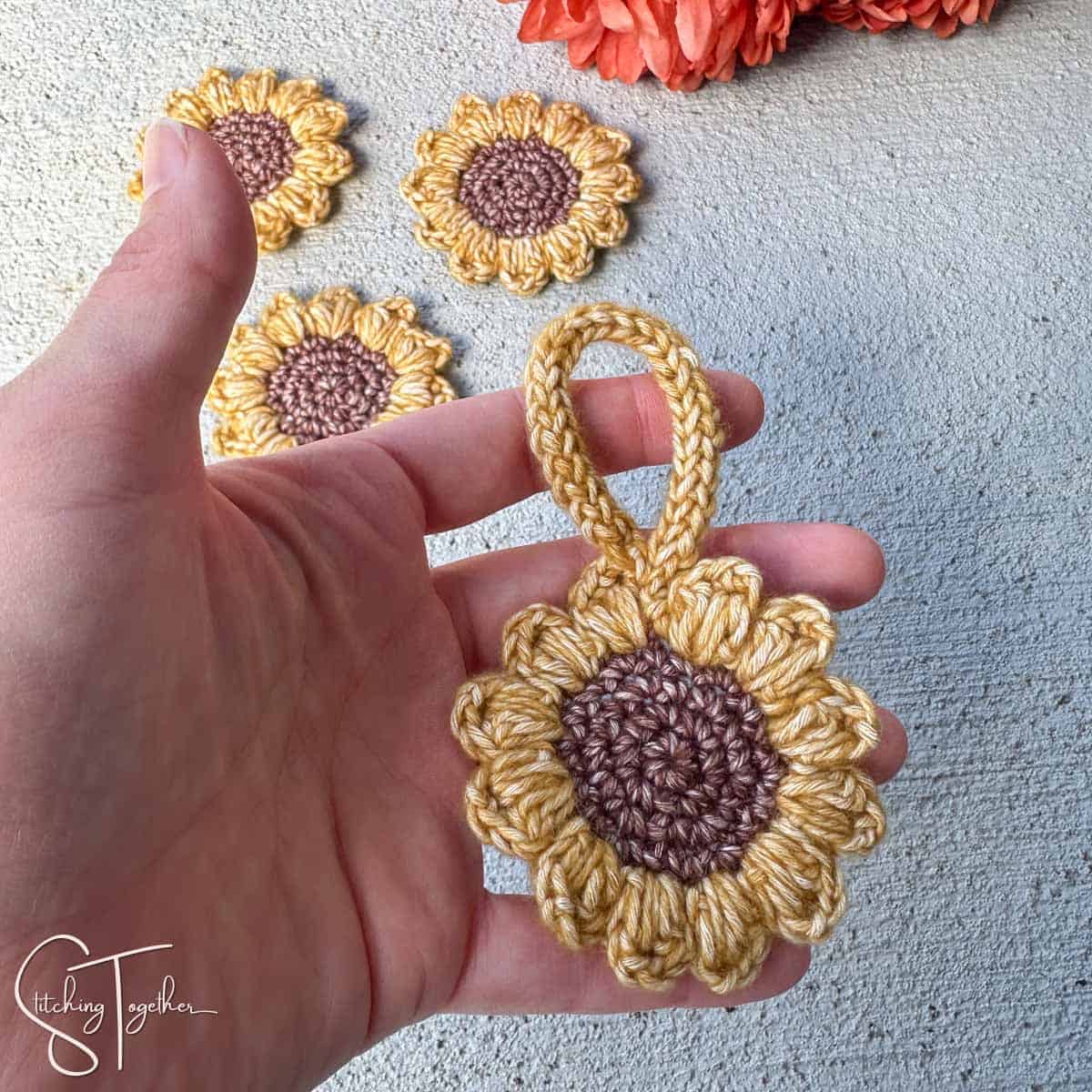 a hand holding a crochet sunflower with a loop with other small sunflowers in the background