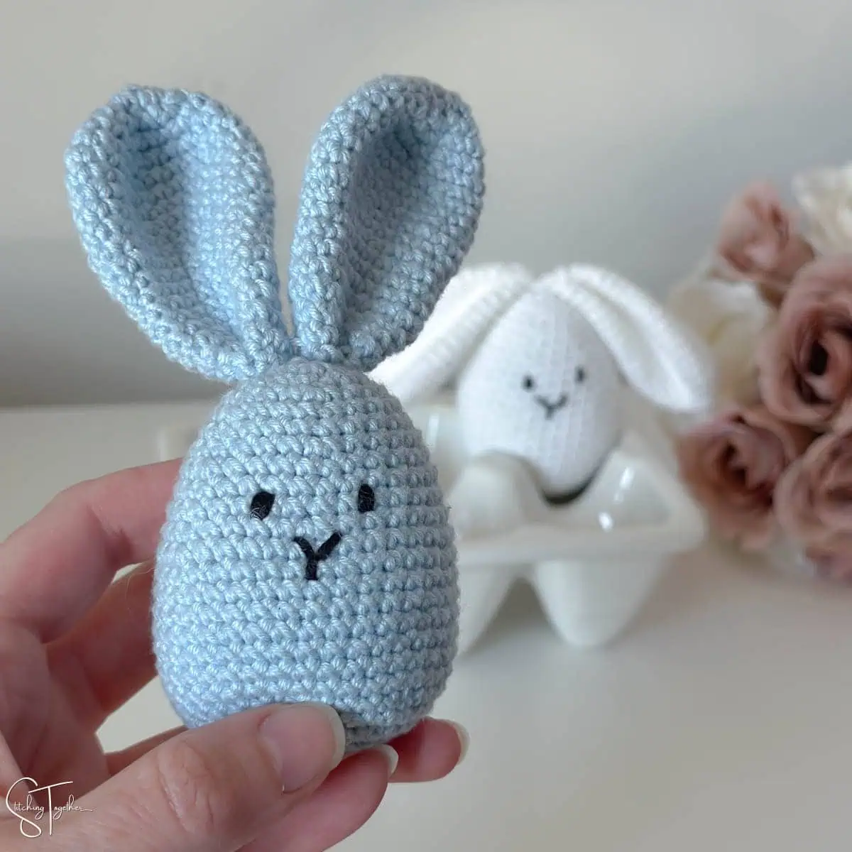 hand holding a crochet bunny egg with another bunny blurred in the background